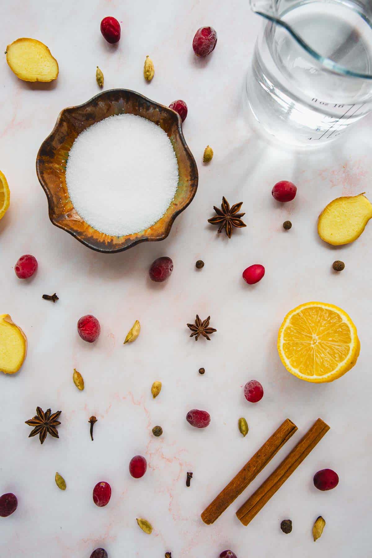 A top view showing various ingredients spread on a light surface, including a bowl of sugar, water in a glass jug, ginger slices, star anise, cranberries, cardamom pods, cloves, cinnamon sticks, and half a sliced orange.