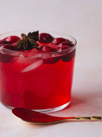 A glass of red beverage with ice cubes, cherries, and a star anise garnish sits on a light-colored surface. Beside the glass is a twisted gold cocktail spoon.