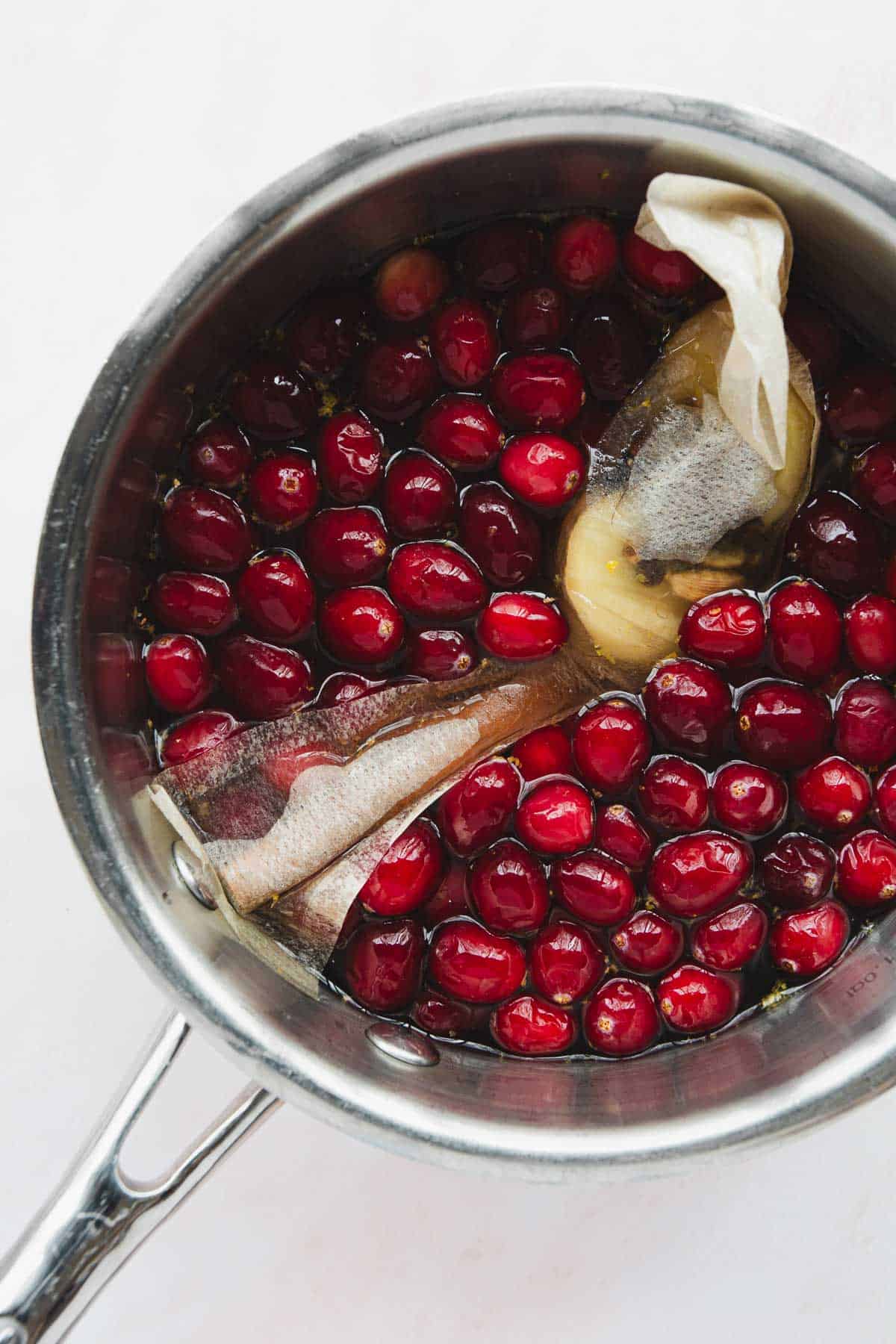 A metal pot filled with fresh cranberries, a peeled piece of ginger, and a tea bag. The ingredients are submerged in a liquid, likely simmering. The pot handle is visible on the left side.