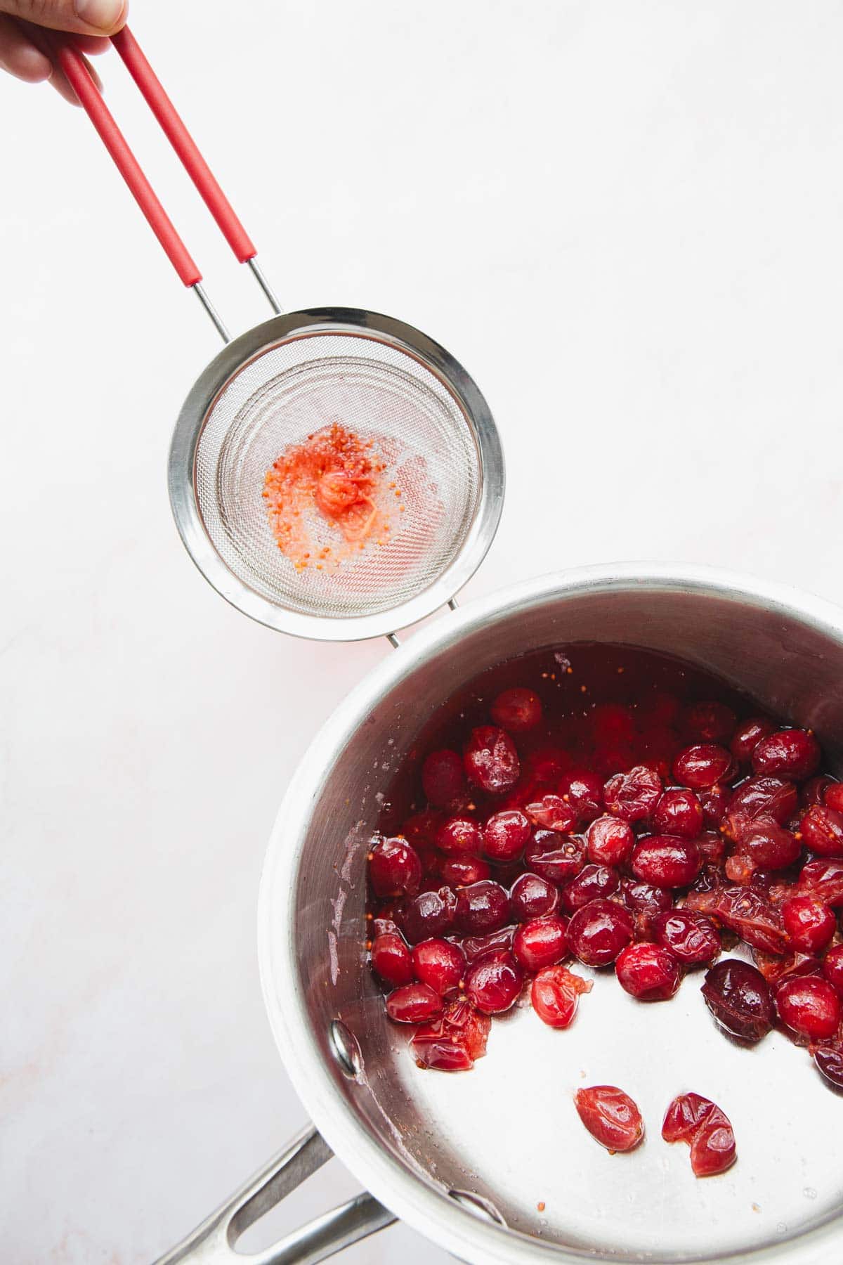 A stainless steel saucepan containing cooked cranberries is placed on a white surface. Above the saucepan, a hand holds a small metal sieve with red residues, likely from straining the cranberries. Red-handled utensils are visible.