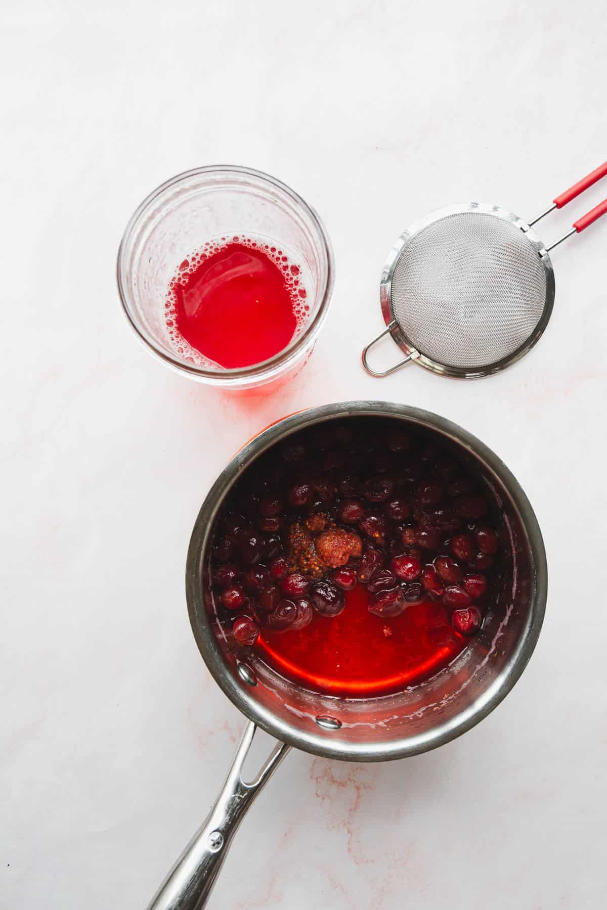 A saucepan filled with a cherry mixture and a cup containing cherry juice are placed on a light-colored surface. A small metal strainer with red handles rests beside the saucepan.
