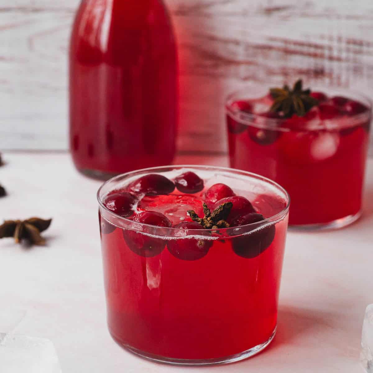 Two glasses of iced spiced cranberry juice with cranberries on top stand on a light surface with a bottle of the same drink in the background. Star anise pods are scattered nearby.