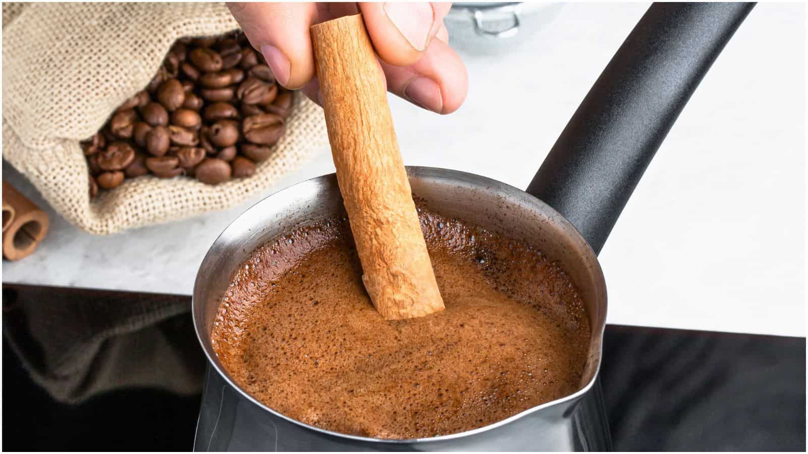 A hand is holding a cinnamon stick and stirring frothed coffee in a small metal pot on a stovetop. A burlap sack with coffee beans is visible in the background. The scene suggests the process of making or flavoring coffee with spices for an enhanced taste.