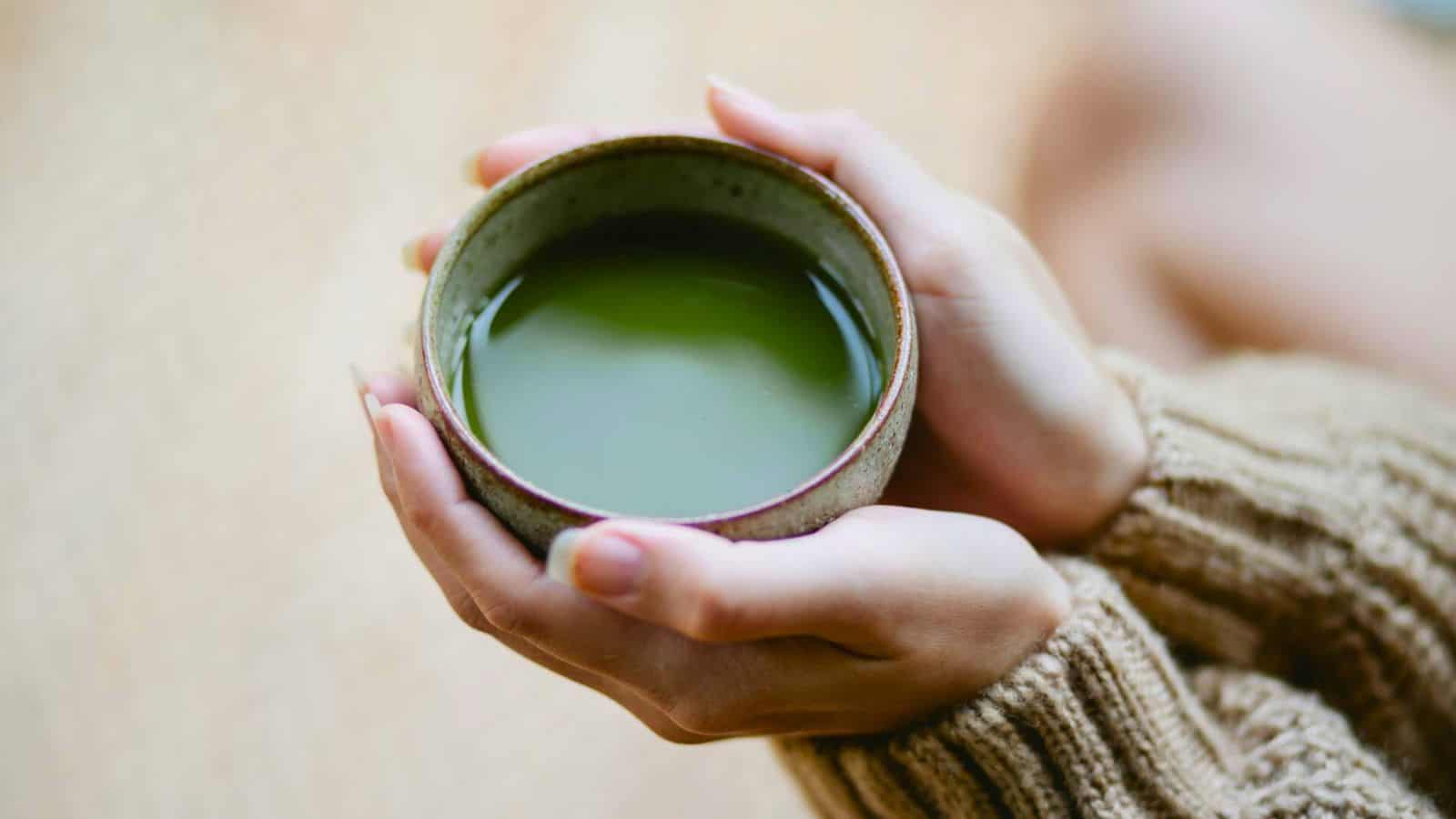 Two hands holding a bowl of matcha. The person is wearing a brown knit sweater. The image is taken close-up, focusing on the bowl and hands, with a soft, neutral background.