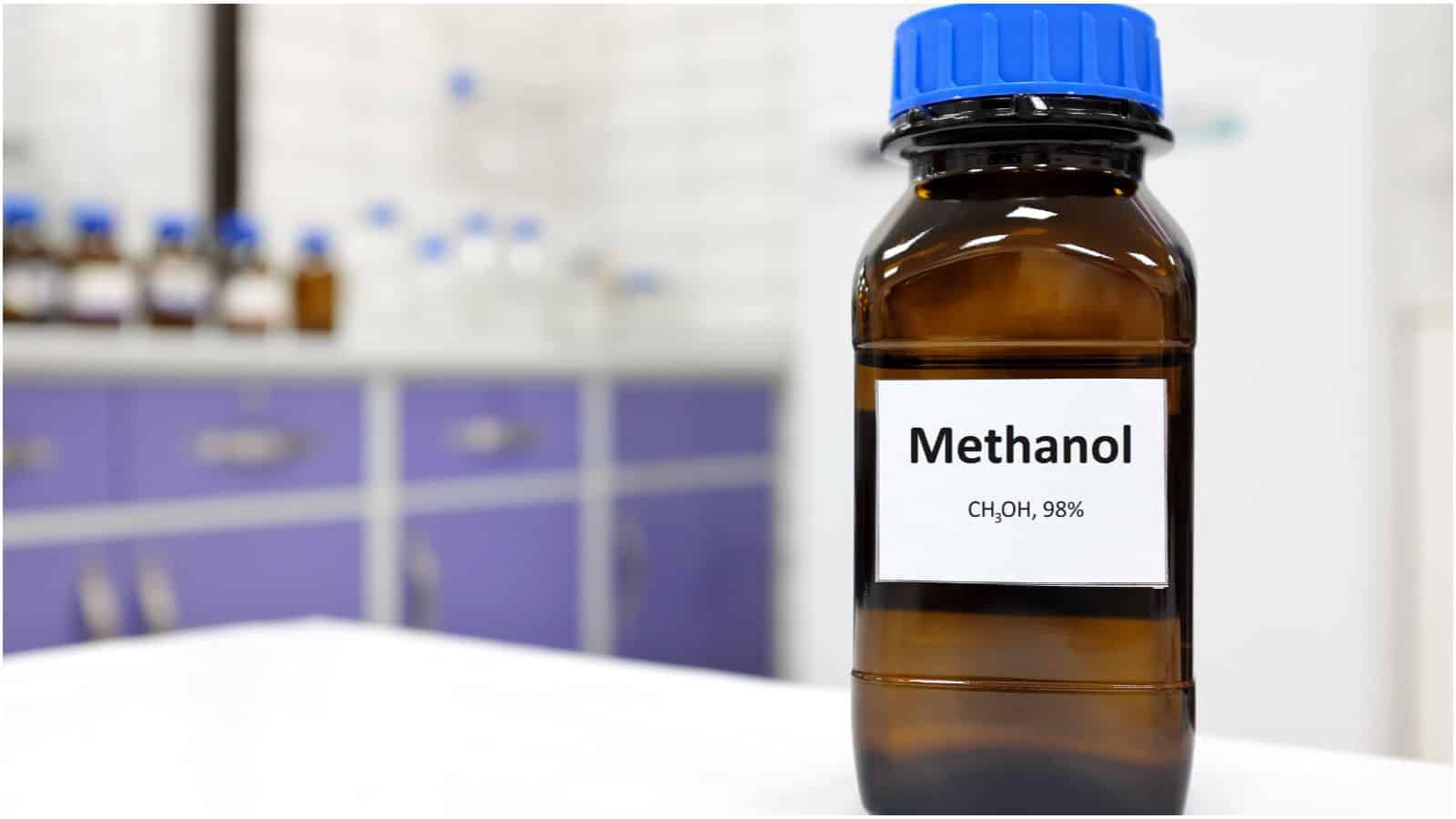 A brown glass bottle with a blue cap labeled "Methanol" and "CH3OH, 98%" stands on a white surface. In the background, blurred laboratory equipment and cabinets with purple doors hint at the types of alcohol you shouldn't be drinking.