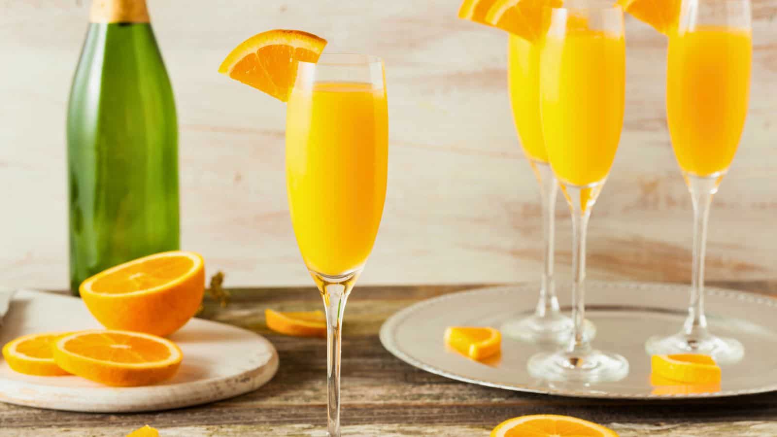 Four glasses of orange mimosa cocktails, which are among the alcoholic drinks that are bad for your teeth, are garnished with orange slices and placed on a wooden table. A green bottle is in the background, and sliced oranges are on a cutting board and plate nearby.