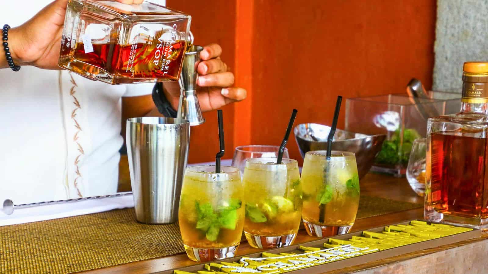 A person is pouring a brown liquid from a bottle labeled "RUM" into a metal shaker. Three glasses with straws and lime wedges, containing an alcoholic drink that can be bad for your teeth, are on the counter. The setting appears to be a bar with other bottles and ingredients in the background.
