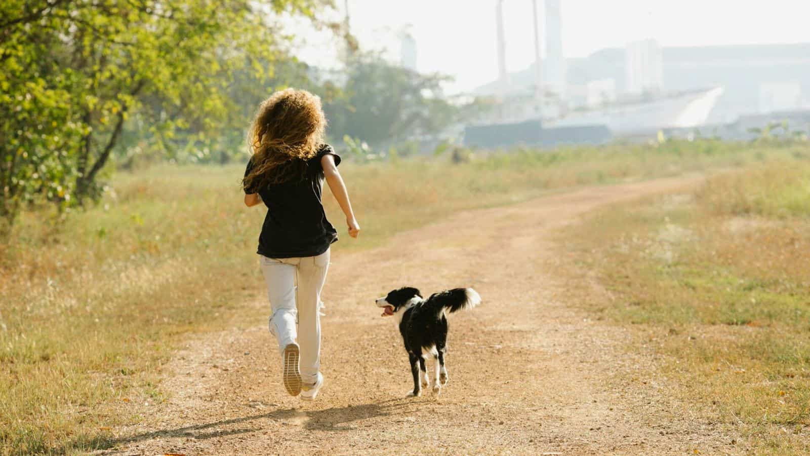 A person with long curly hair wearing a black shirt and light pants is running on an unpaved path in a grassy, open area, promoting a healthier lifestyle to stop drinking alcohol. A black and white dog runs beside them. In the background, there are trees and the outline of industrial buildings.