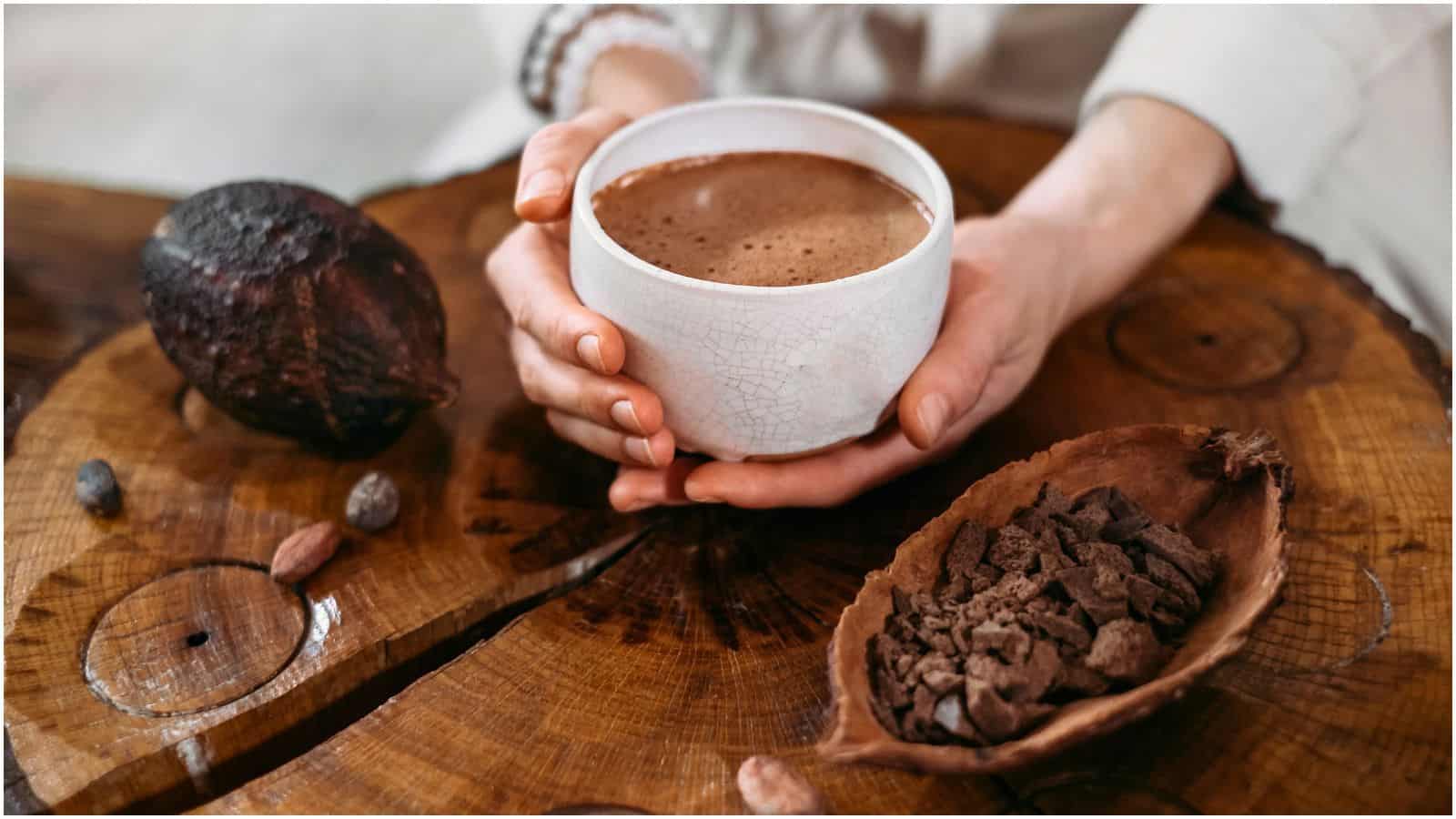 A pair of hands holding a white ceramic cup filled with a frothy chocolate drink on a wooden surface. There is a halved cacao fruit and some cacao nibs beside the cup.