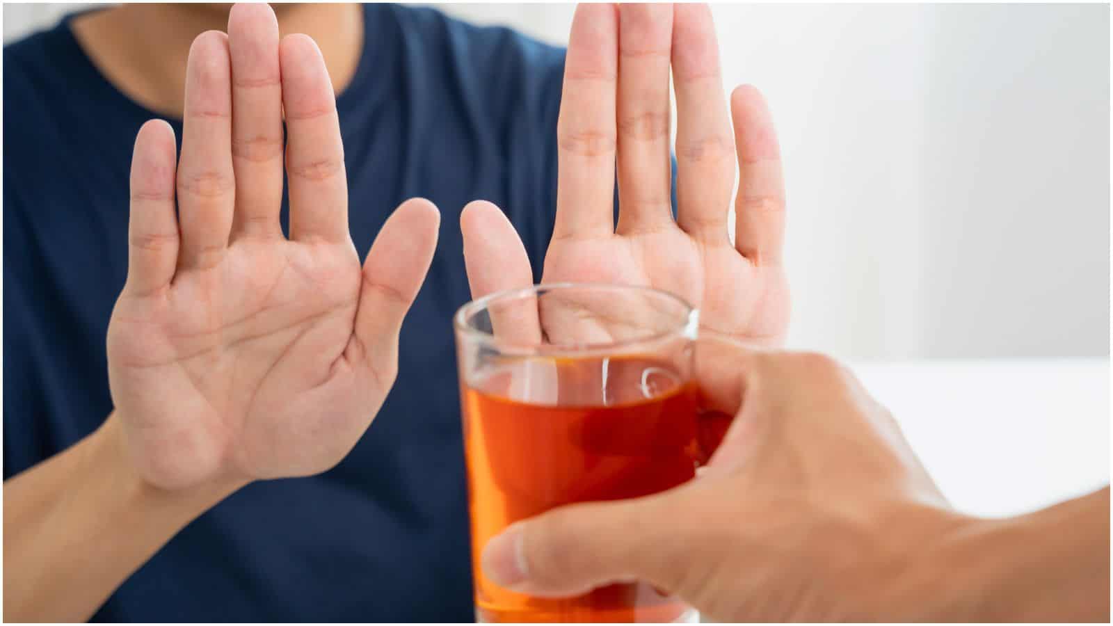 A person in a dark blue shirt holds both hands up, palms outward, in a gesture of refusal. Another hand, extending from outside the frame, offers a glass of orange liquid to the person, who appears to be declining the drink, signaling their decision to stop drinking alcohol.