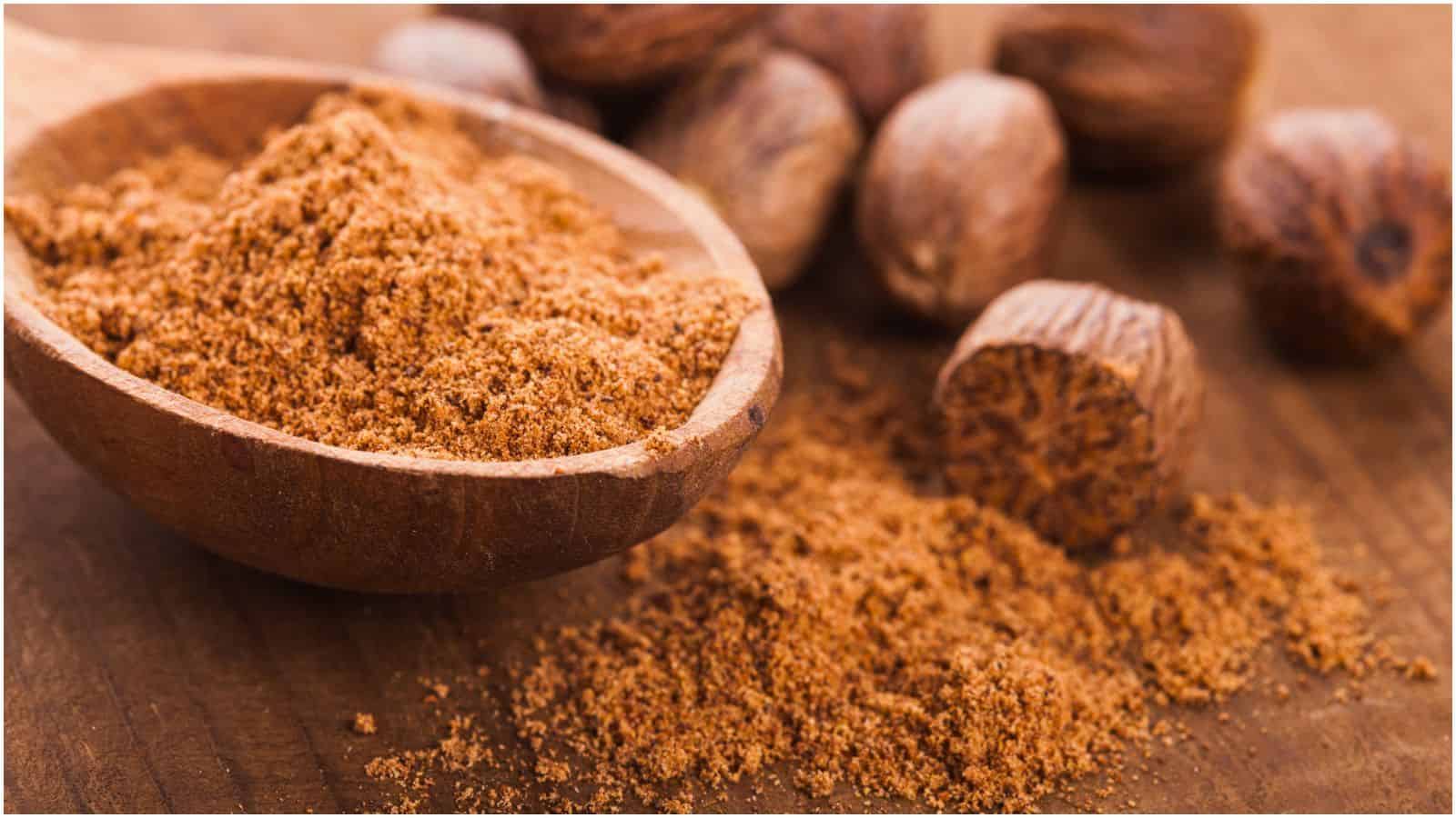 A wooden spoon filled with ground nutmeg, one of the perfect spices for coffee, sits on a wooden surface. Loose ground nutmeg is scattered beside the spoon, with whole nutmeg seeds visible in the background.