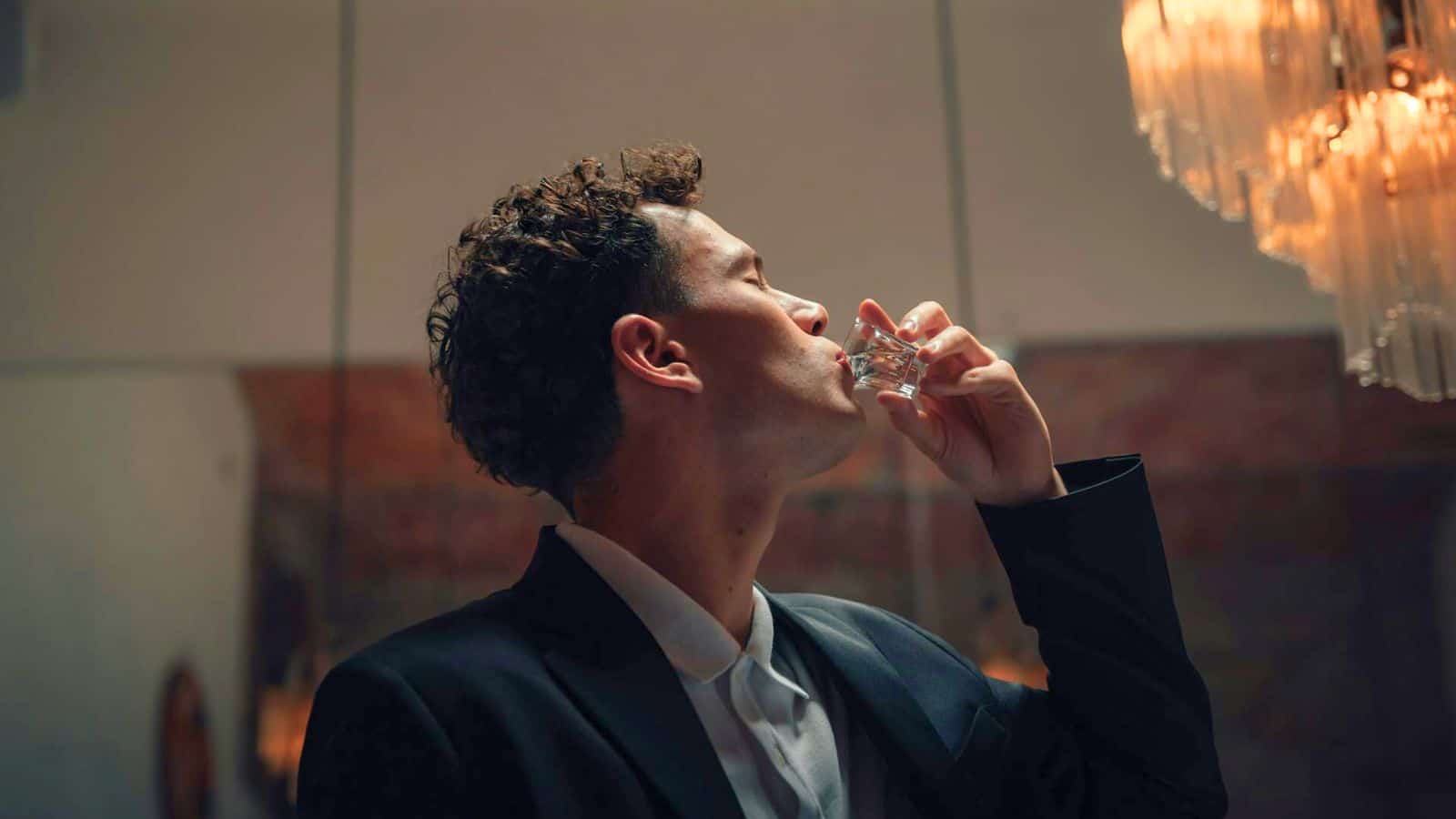 A person with curly hair, dressed in a dark suit, is drinking from a small glass of alcohol. They tilt their head back while standing under a chandelier. The background is softly lit with warm tones.