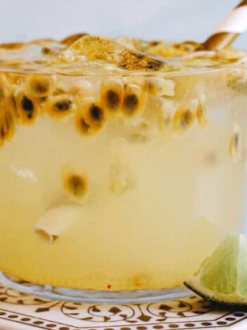 A glass filled with a light yellow drink, containing ice and passion fruit seeds, sits on a decorative coaster. A striped paper straw is placed in the drink, and a small lime wedge is positioned at the base of the glass. The background is light blue.