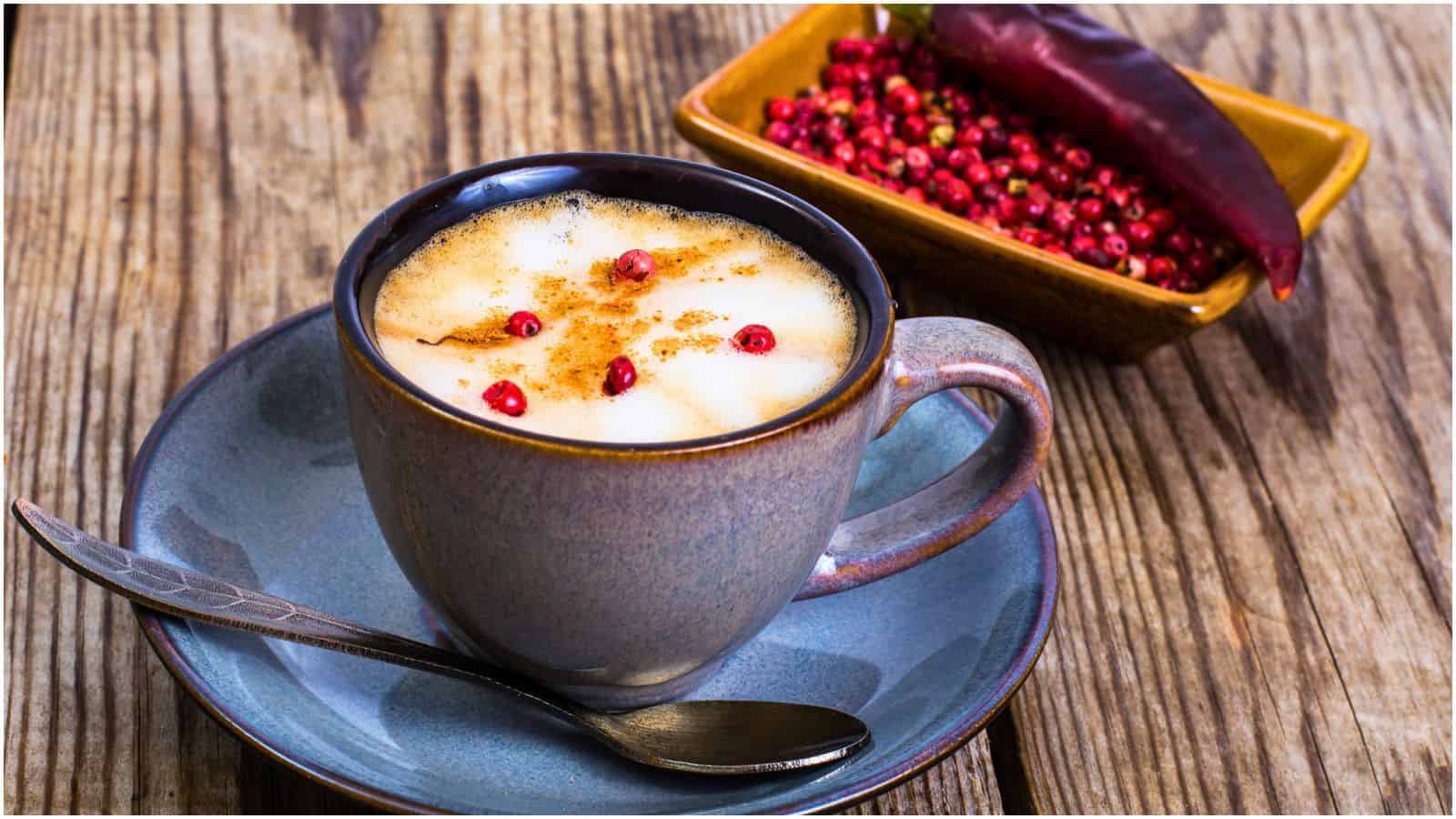 A ceramic cup filled with frothy coffee topped with red pepper flakes is on a matching saucer with a teaspoon. Next to it is a rectangular dish containing spices for coffee: red peppercorns and a whole red chili pepper. The setup is on a wooden surface.