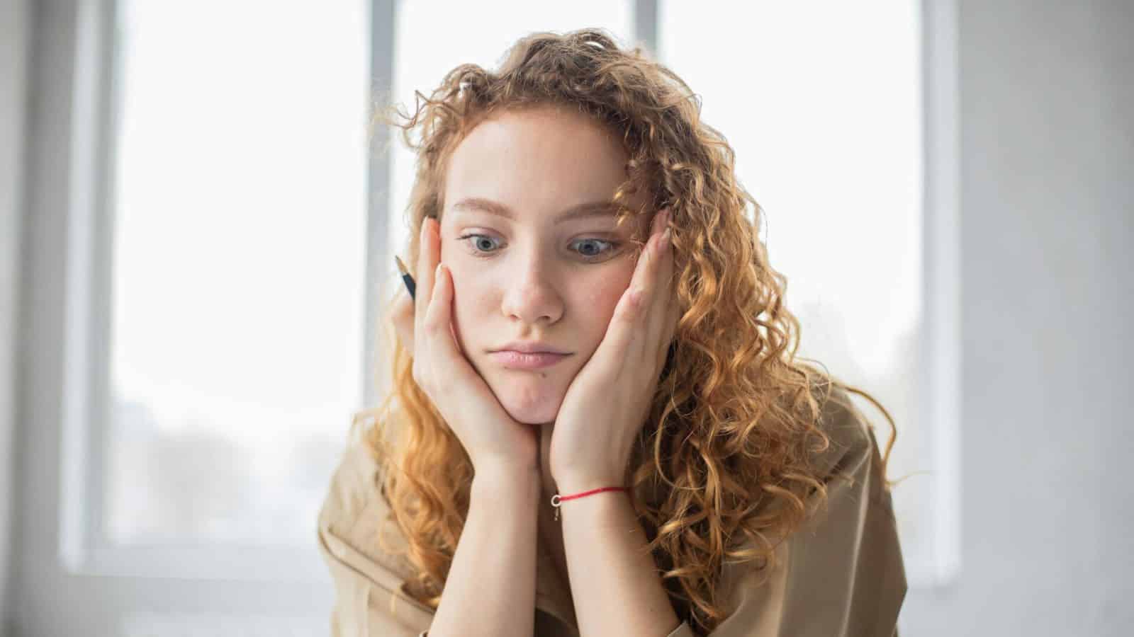 A person with curly hair is resting their face in their hands while looking downward with a thoughtful expression. They are seated indoors, in front of a large, bright window, perhaps contemplating the complexities of alcohol rules.