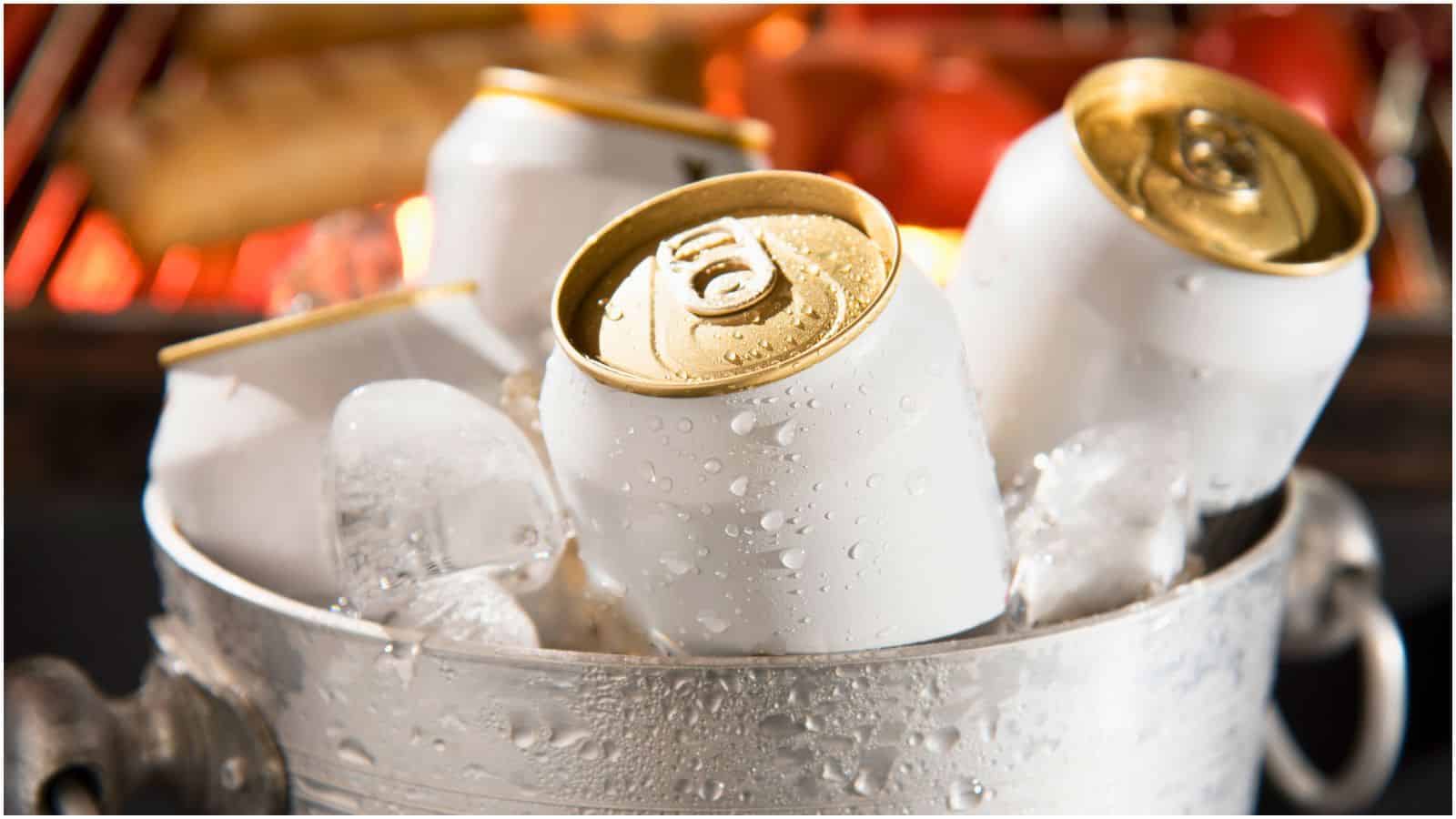 Four white aluminum cans with gold tops are chilled in an ice-filled metal bucket. The background is slightly blurred, showing a warm, orange glow from a nearby heat source like a grill or fire—a subtle reminder to be mindful of the types of alcohol you shouldn't be drinking.