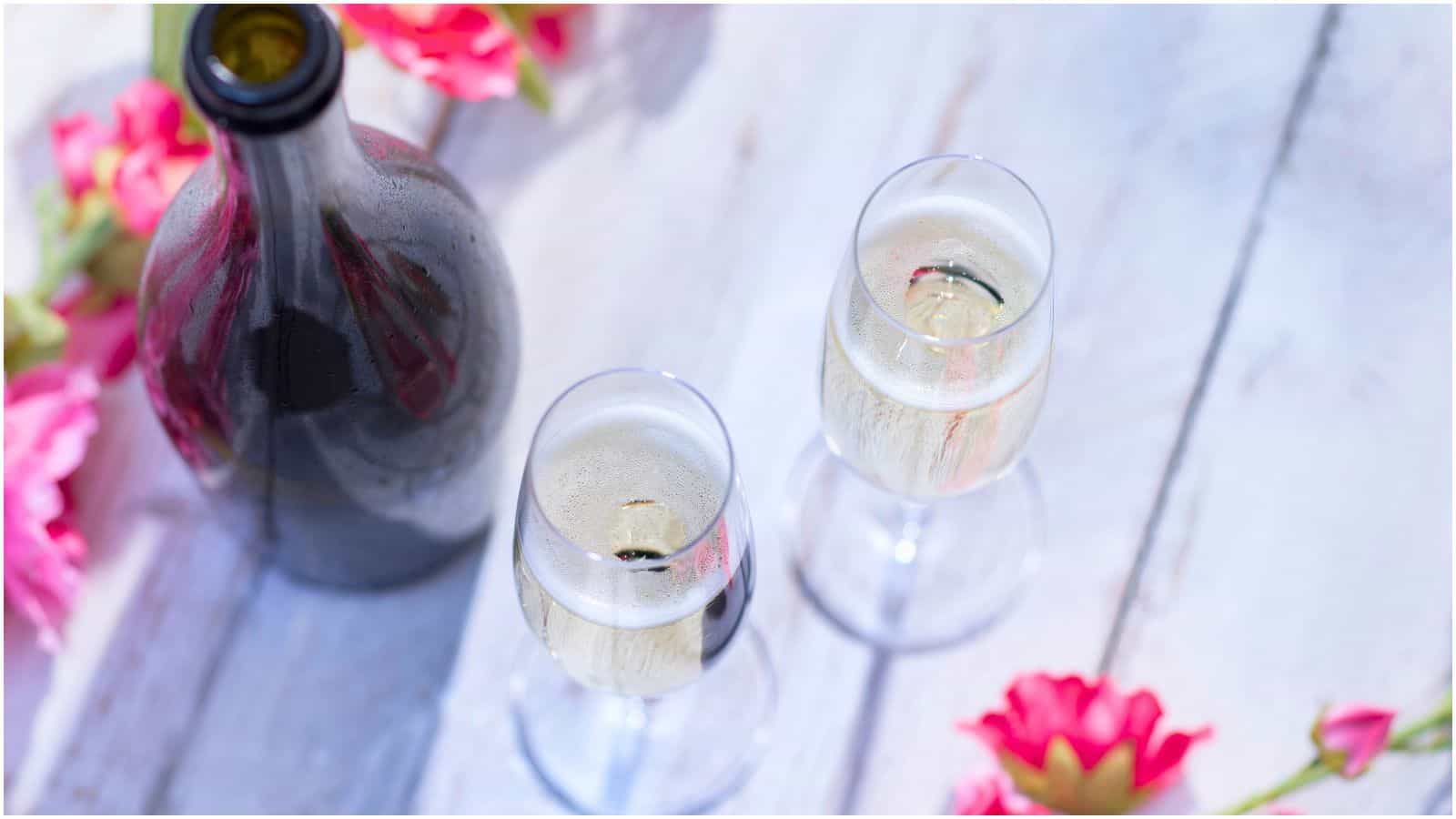 A bottle of sparkling wine is placed on a white wooden surface, accompanied by two filled wine glasses. Pink flowers are scattered around the scene. While the composition suggests a celebratory or romantic setting, it's worth noting that alcoholic drinks like these can be bad for your teeth.