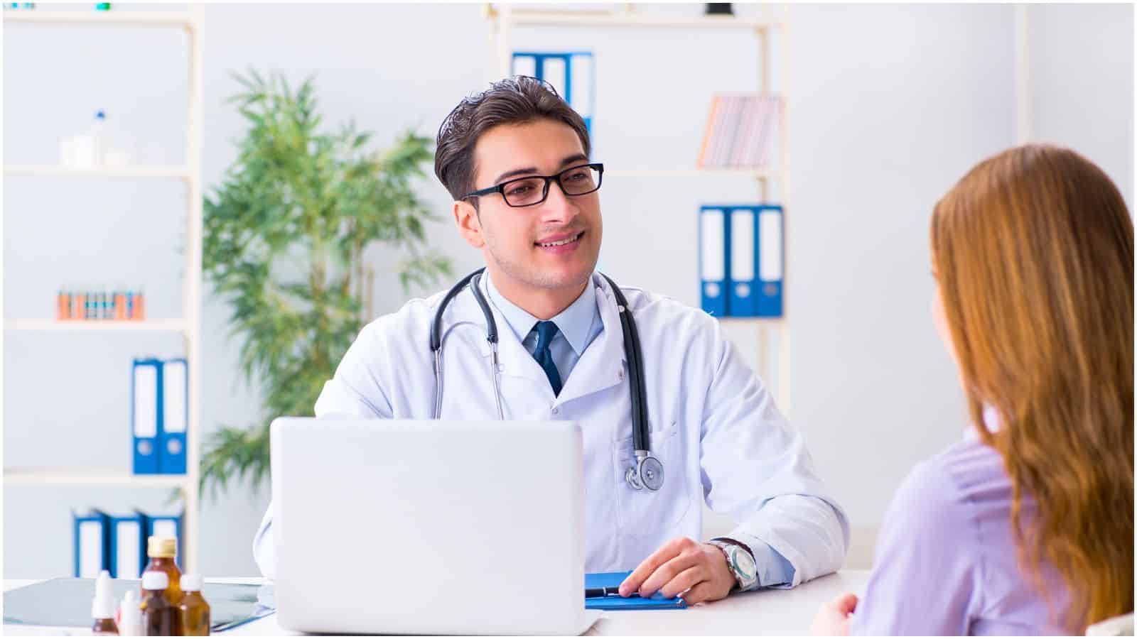 A doctor wearing a white coat and stethoscope, sitting at a desk with a laptop, smiles at a patient who is sitting across from him. Amidst the bookshelves with files, a plant, and some medicine bottles on the desk, he gently advises the patient to stop drinking alcohol for better health.