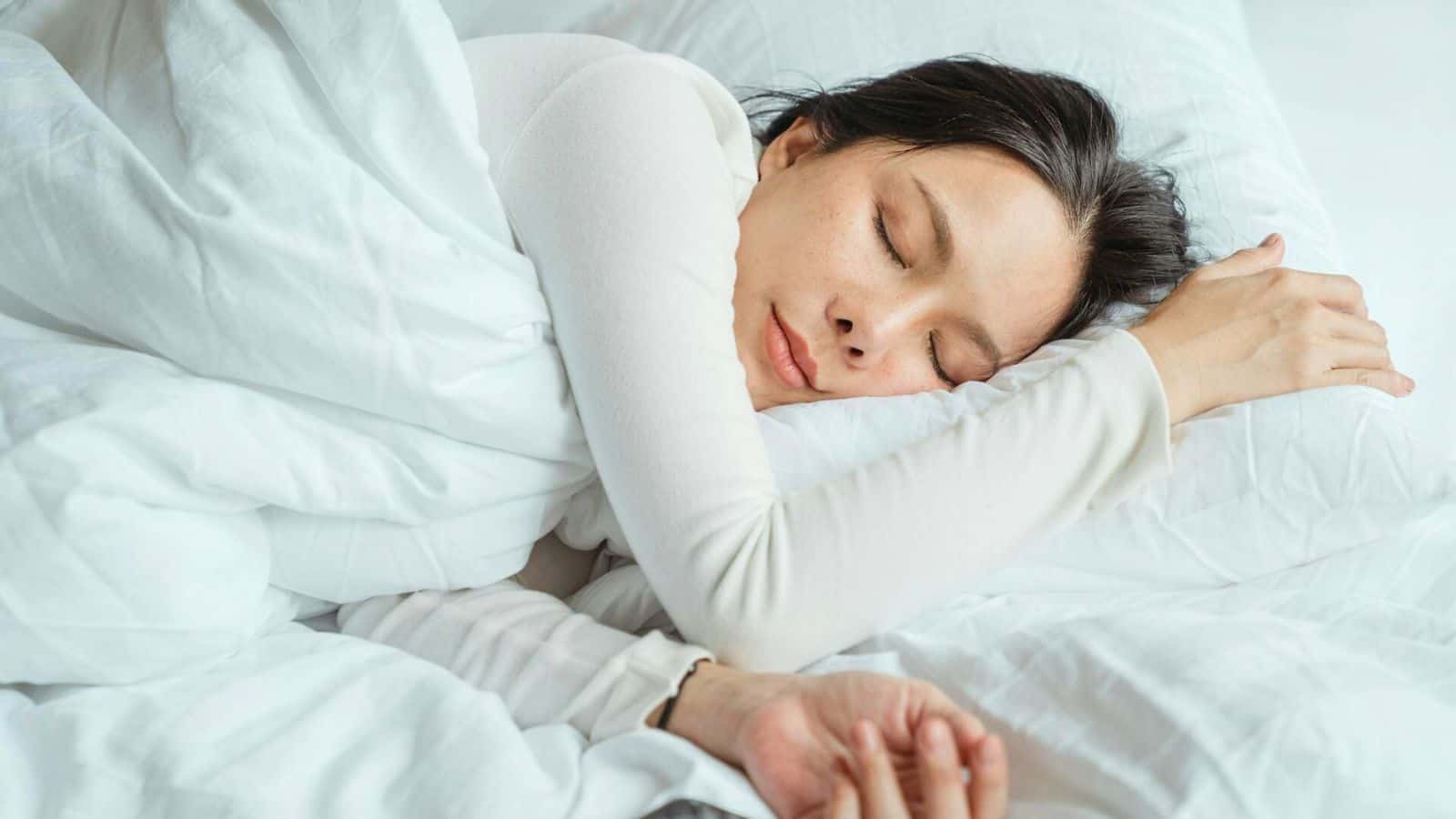 A person wearing a white long-sleeve shirt is sleeping peacefully on their side with their head resting on a white pillow and their hand under their face. The bed is covered with a white duvet, suggesting they've enjoyed a soothing glass of lemon water before bed for better rest.