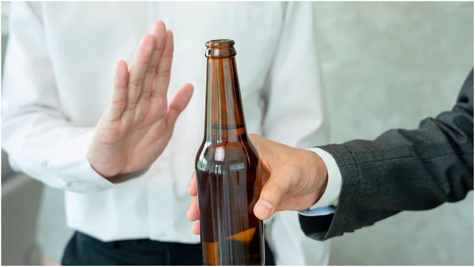 A person in a white shirt holds up their hand in a gesture of refusal as another individual in a dark suit offers a brown glass bottle. The image suggests someone declining a drink, adhering to personal alcohol rules.