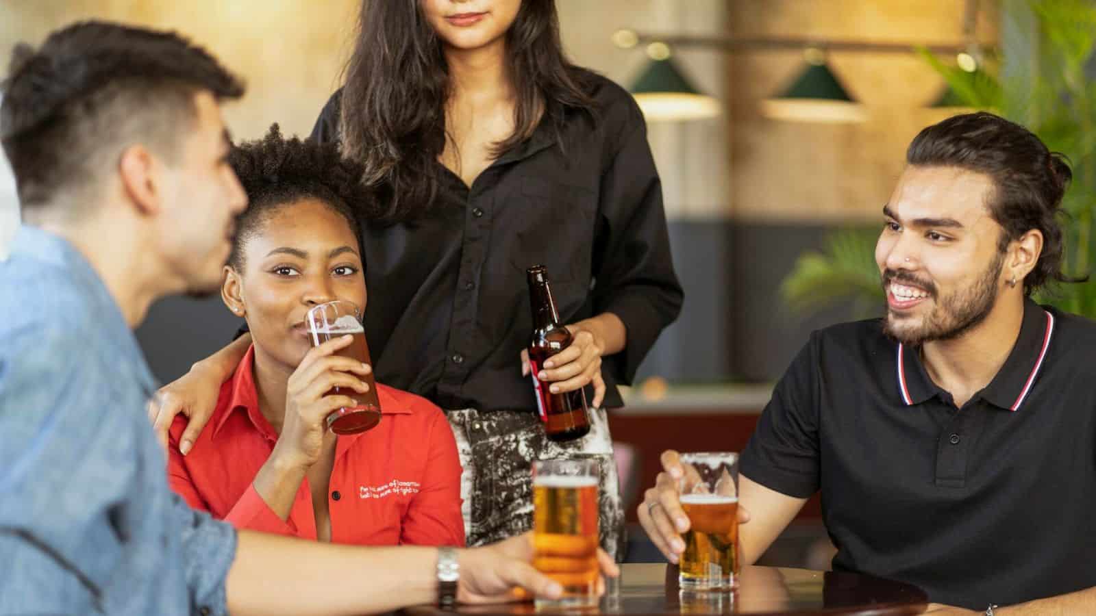 Four people are gathered around a table in a bar setting. Two of them are seated, each holding a pint of beer and smiling, while a third person stands with a beer bottle. Despite the casual and relaxed atmosphere, they seem aware of the alcohol rules in place.