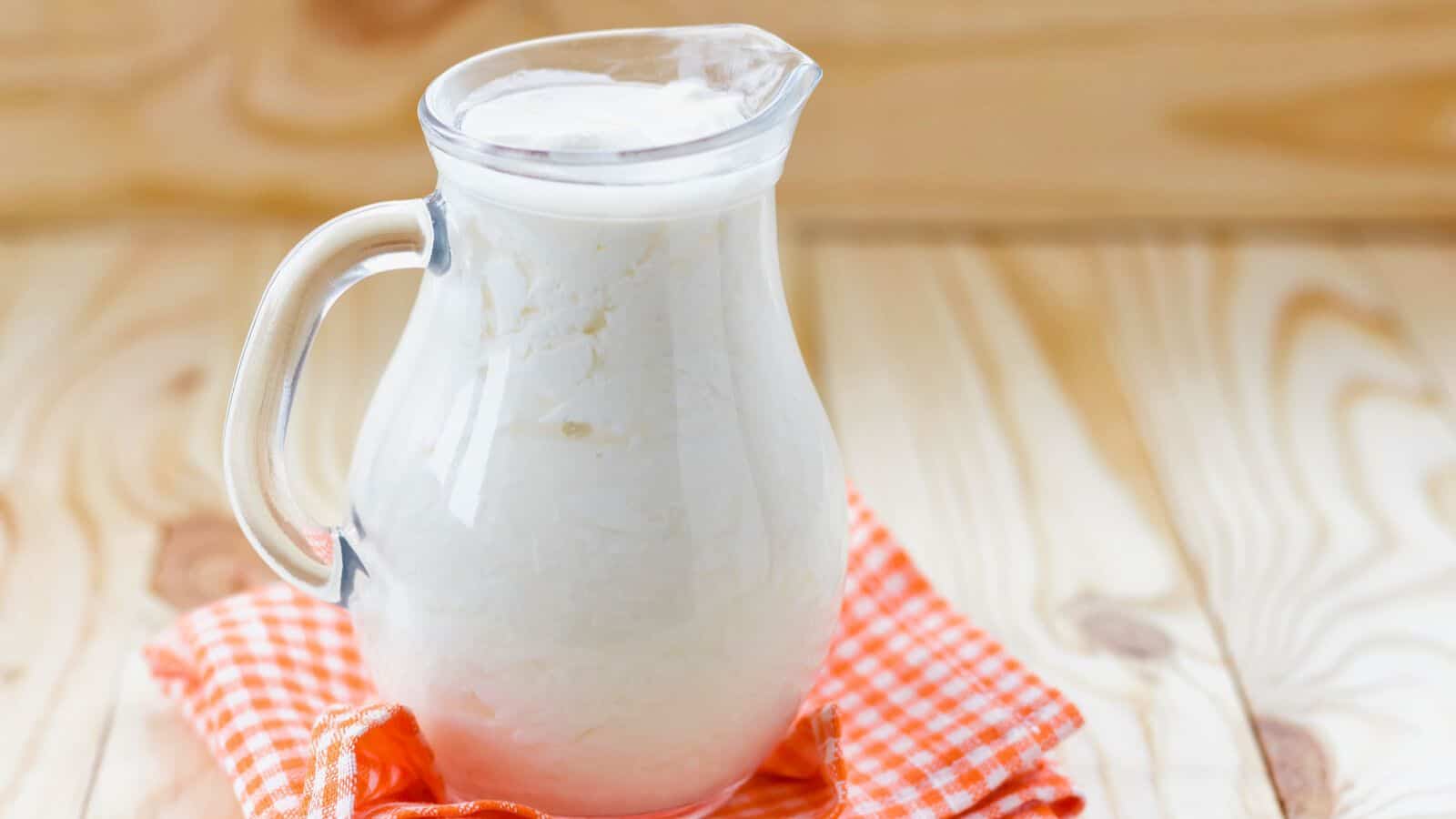 A clear glass pitcher filled with a thick white liquid, likely kefir, is placed on an orange checkered cloth. The background consists of a light wooden surface with visible grain patterns.