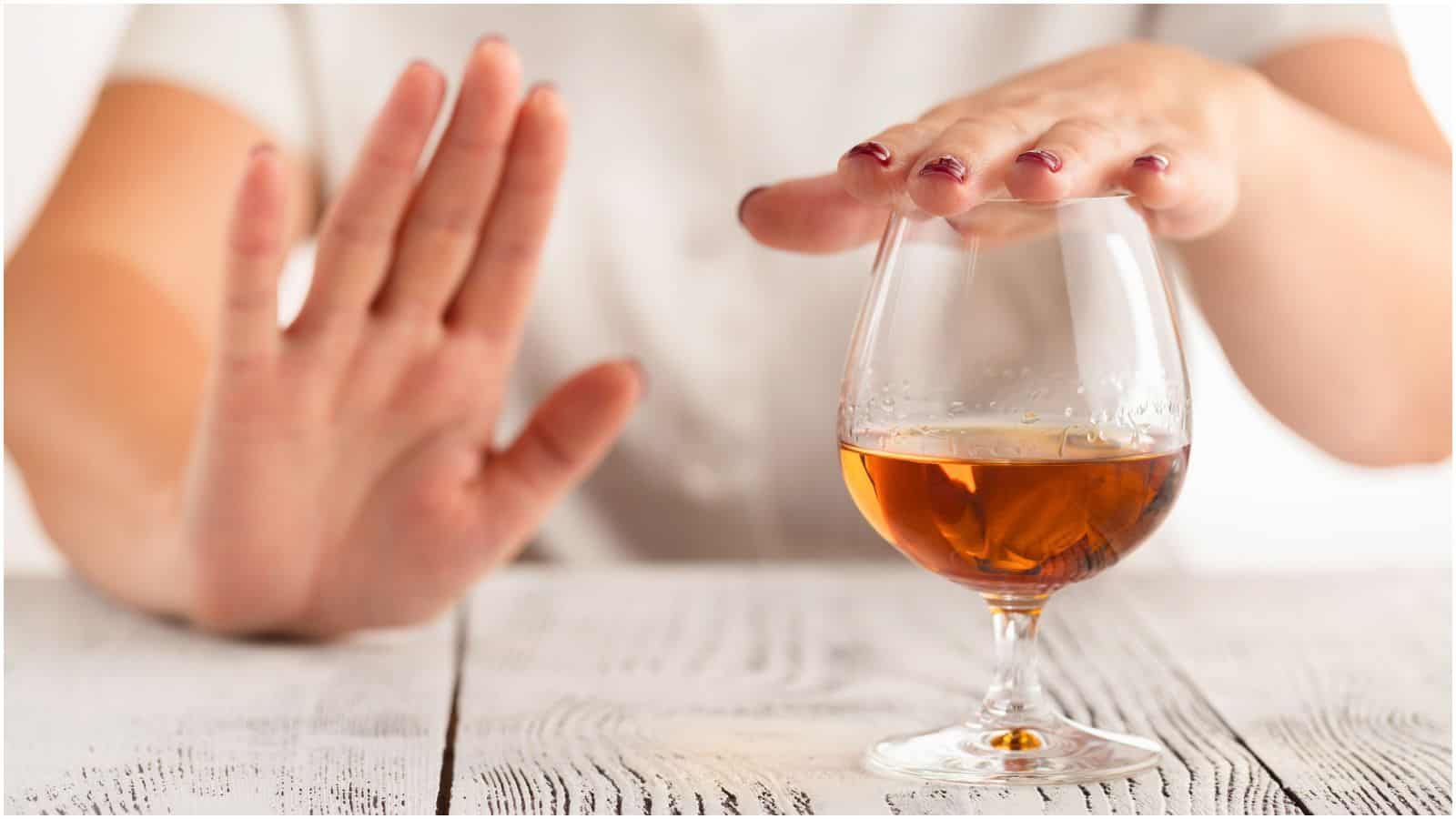 A person is making a stop gesture with one hand while placing the other hand over a glass of amber-colored liquor on a wooden surface, suggesting they are refusing or avoiding types of alcohol you shouldn't be drinking.