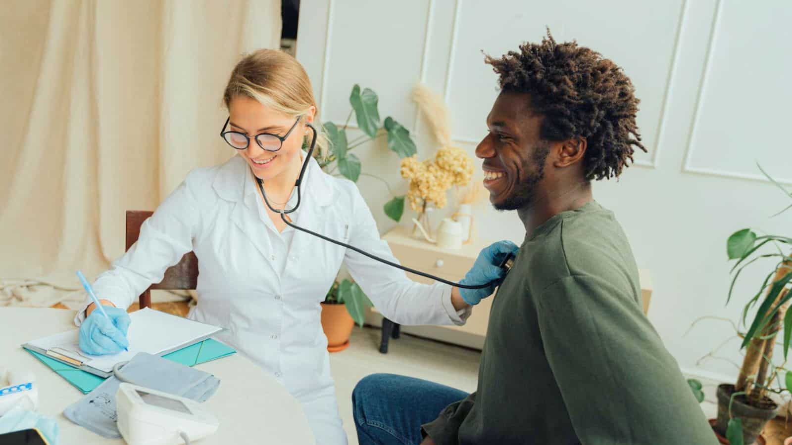 A healthcare professional in a white coat and gloves uses a stethoscope to examine a smiling individual sitting next to her. She is writing notes on a clipboard, possibly about the patient's list of sugary drinks. They are in a room with plants and medical equipment.