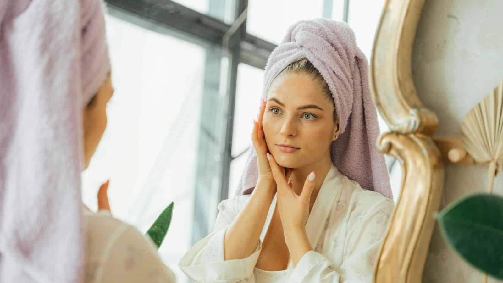 A person with a purple towel wrapped around their head stands in front of a mirror, touching their face with both hands. They are wearing a white robe and have a focused expression, perhaps thinking about the benefits of lemon water before bed. A plant and a decorative mirror frame are visible in the background.