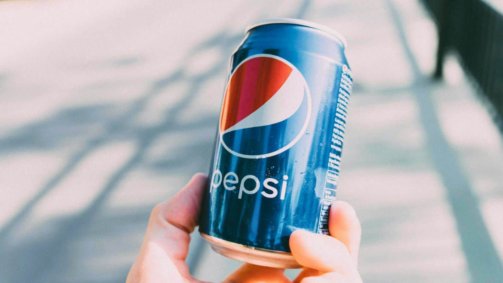 A hand is holding a blue Pepsi can with the Pepsi logo and white text, a classic choice from the list of sugary drinks. The background is out of focus, featuring a bridge railing in the sunlight.