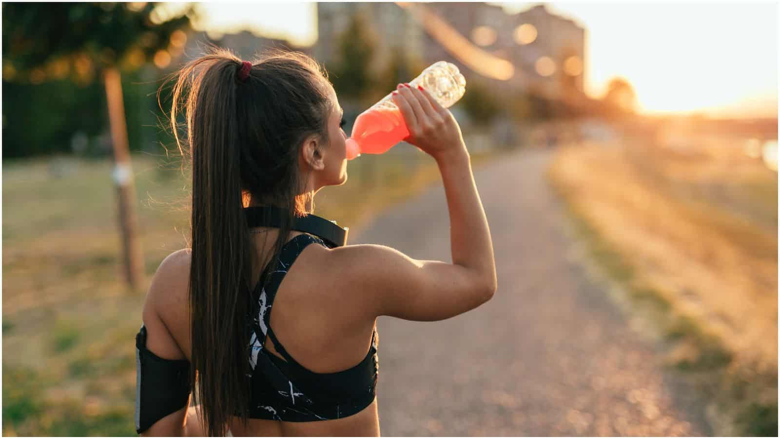 A woman with a ponytail, wearing a black sports top, drinks a pink beverage from a bottle. She is standing on a dirt path with trees and buildings in the background under a setting sun, appearing to enjoy something straight from her list of sugary drinks.