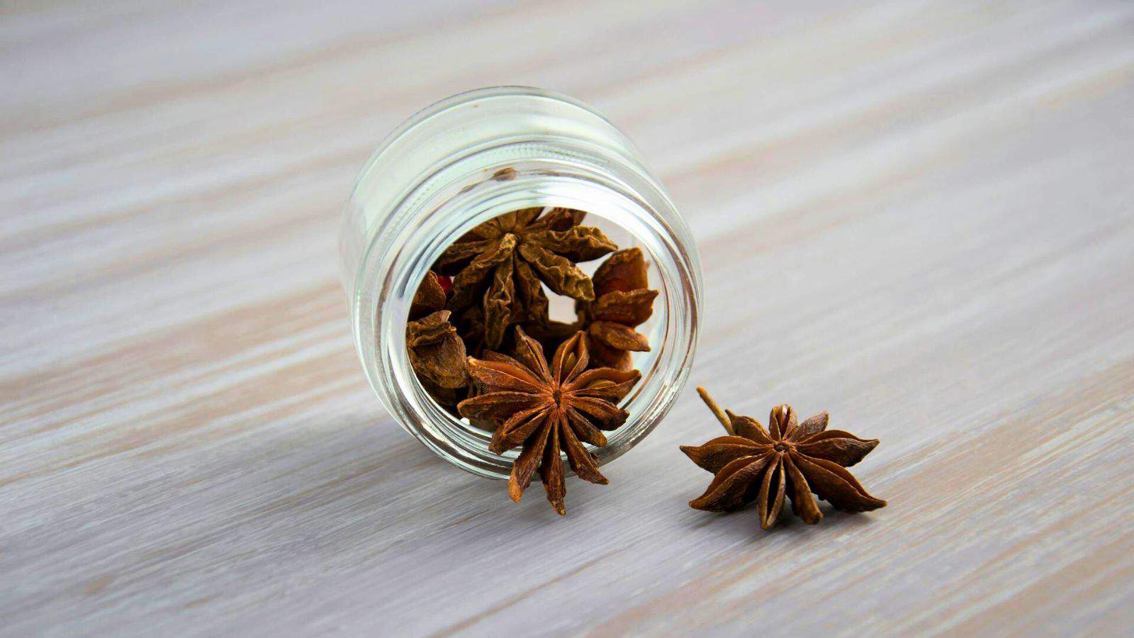 A glass jar on its side with star anise spilling out onto a wooden surface. One star anise is fully outside the jar, while several others, perfect as spices for coffee, are partially inside it.