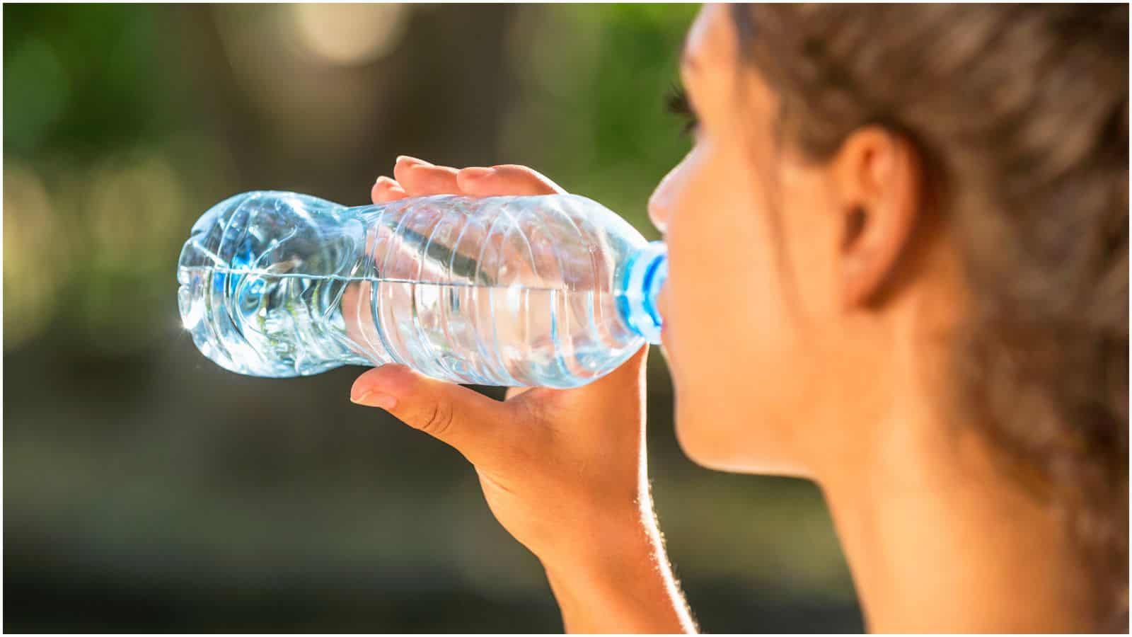 A close-up image of a person holding a clear plastic water bottle and taking a sip. The individual is facing away from the camera, with green, out-of-focus foliage in the background. The setting appears to be outdoors in natural light, making it an ideal moment to reflect on strict alcohol rules in public spaces.