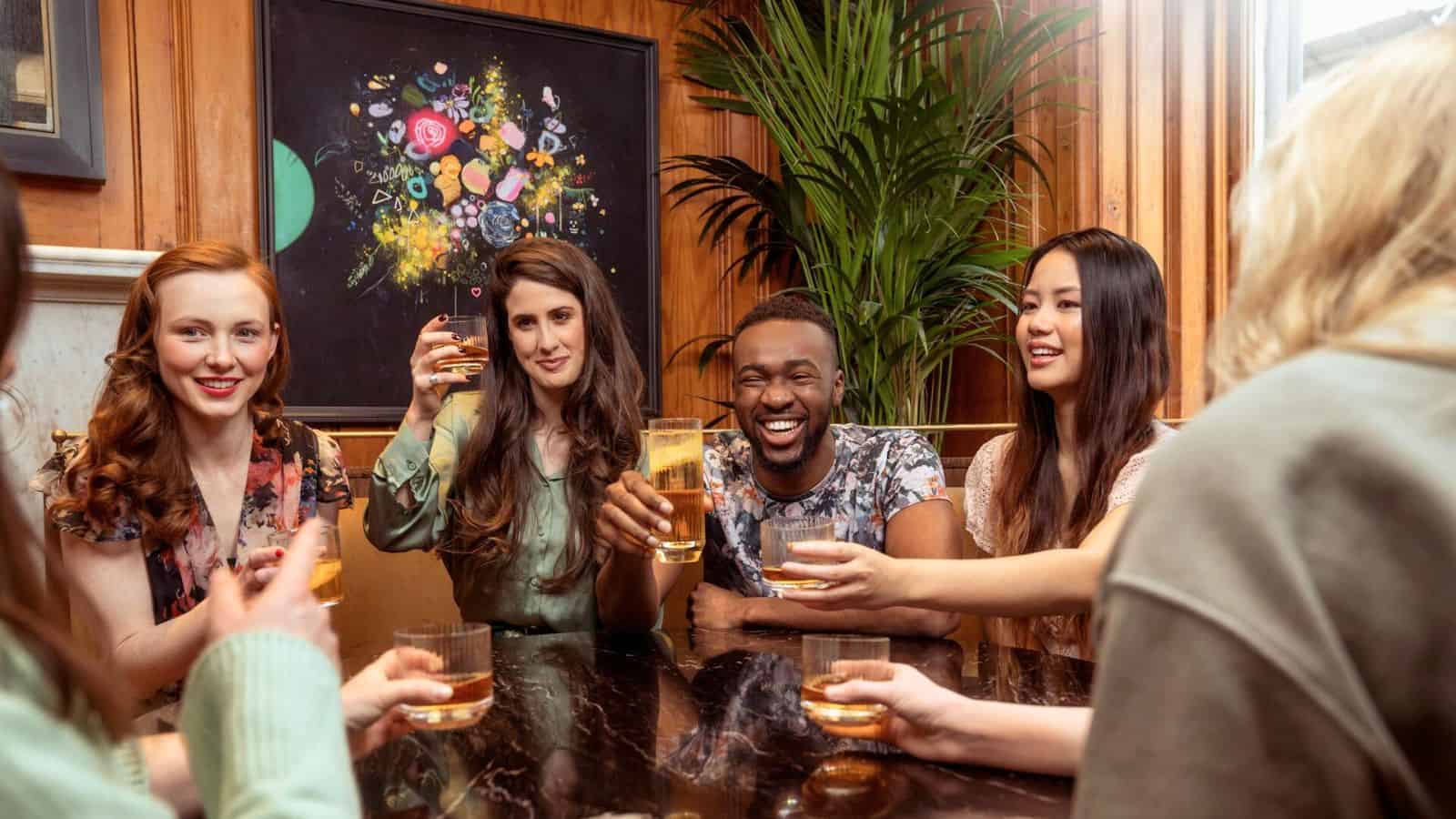 A group of five people are sitting around a dark marble table, clinking glasses and enjoying their drinks while following alcohol rules. They are all smiling, with a vibrant floral painting and a large green plant setting the background.