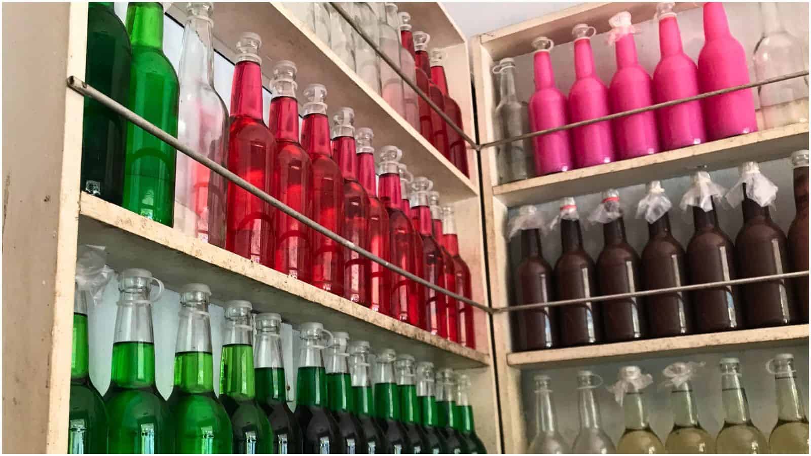Shelves filled with colorful glass bottles, including everything you need to know about simple syrup. The bottles are arranged by color&mdash;green, red, pink, brown, and light-colored liquids. The shelves are separated by metal bars, and some bottles are covered with plastic.