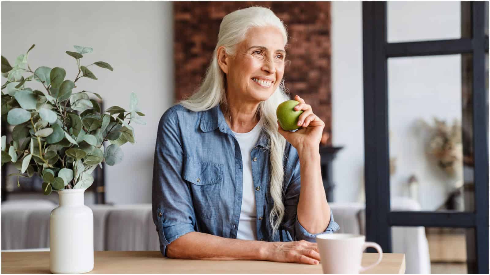 A woman with long white hair is sitting at a table, holding a green apple and smiling. She's wearing a blue shirt over a white top. In front of her are a white mug with tea and a vase with green foliage, reflecting her new lifestyle choice to stop drinking alcohol.