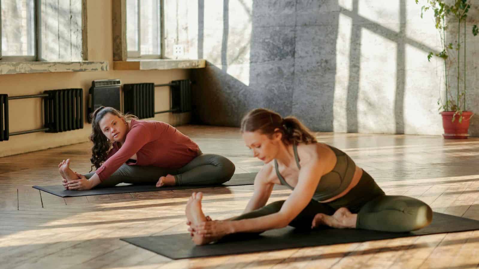 Two individuals are practicing yoga on mats in a well-lit studio with large windows. Both are seated, extending one leg straight out and reaching toward their toes. The studio has wooden flooring and a potted plant in the background.
