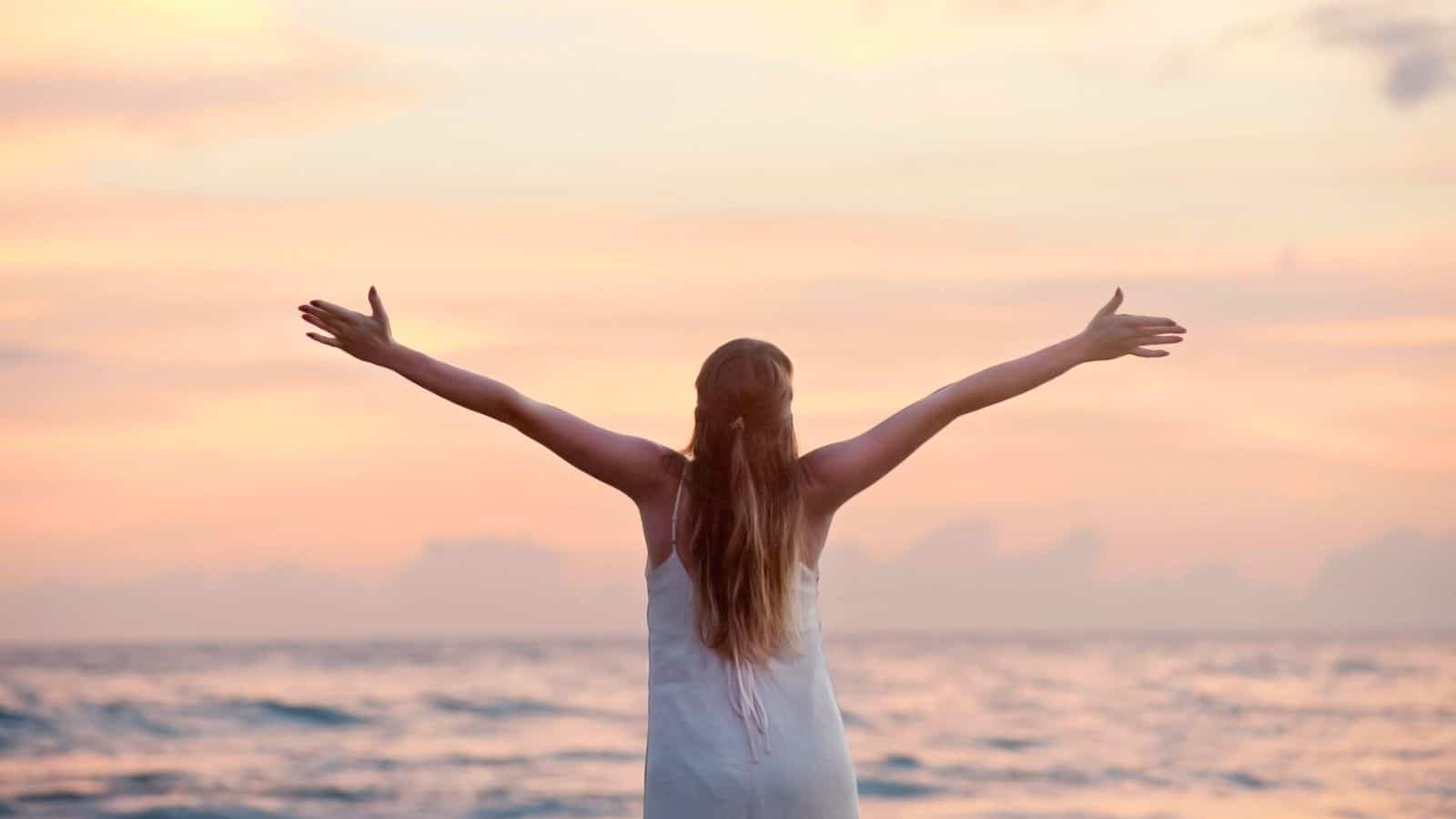 A person with long hair is standing with their back to the camera, arms outstretched, facing the ocean during what appears to be sunset or sunrise. The sky is filled with soft pink and orange hues.