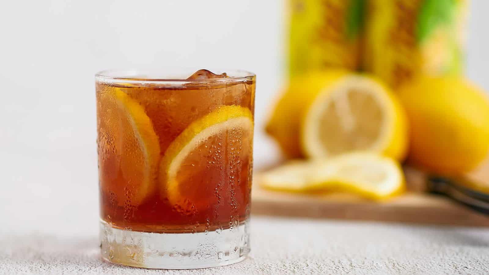 A glass of iced tea with lemon slices inside, condensation on the glass. In the blurred background, there are two lemons, one whole and one sliced, placed on a wooden surface; a refreshing choice amidst a list of sugary drinks.