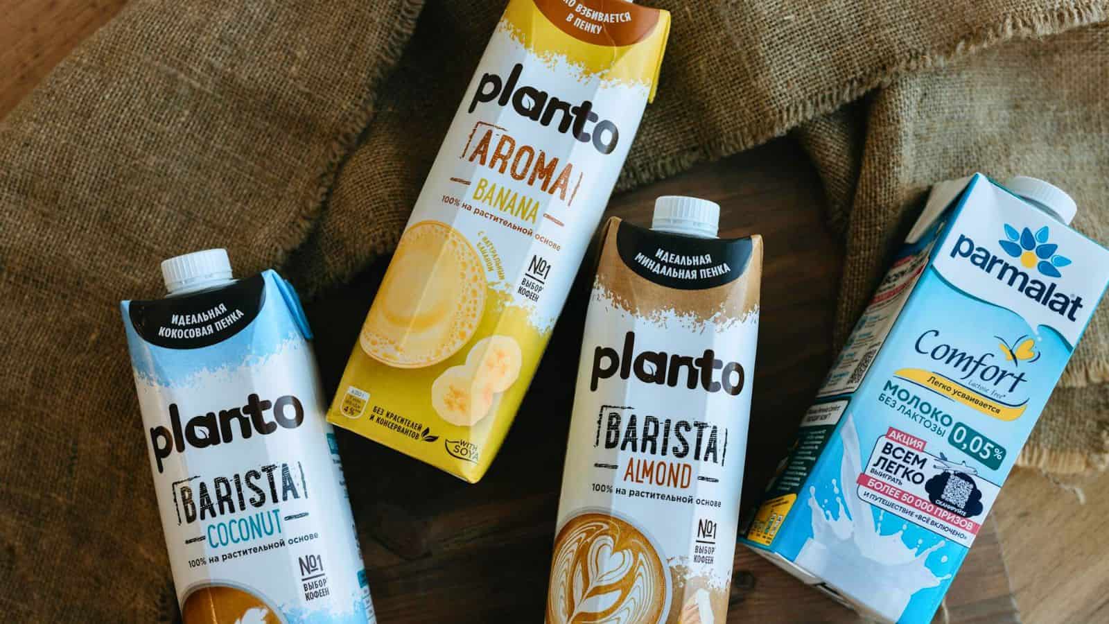 Four cartons of plant-based beverages are displayed on a wooden surface with a burlap background. The brands include "Planto" with flavors like coconut, banana, and almond, and "Parmalat Comfort." Unlike your typical list of sugary drinks, these options provide healthier alternatives without sacrificing taste.