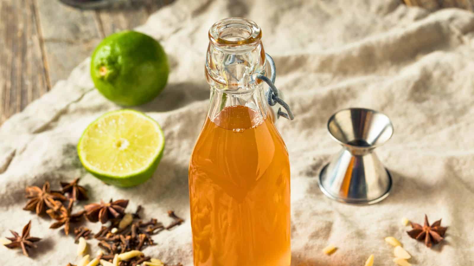 A bottle of orange liquid stands on a cloth. Surrounding it are a halved lime, a whole lime, a metal jigger, and scattered spices including star anise and chopped nuts&mdash;everything you need to know about mixing simple syrup essentials.