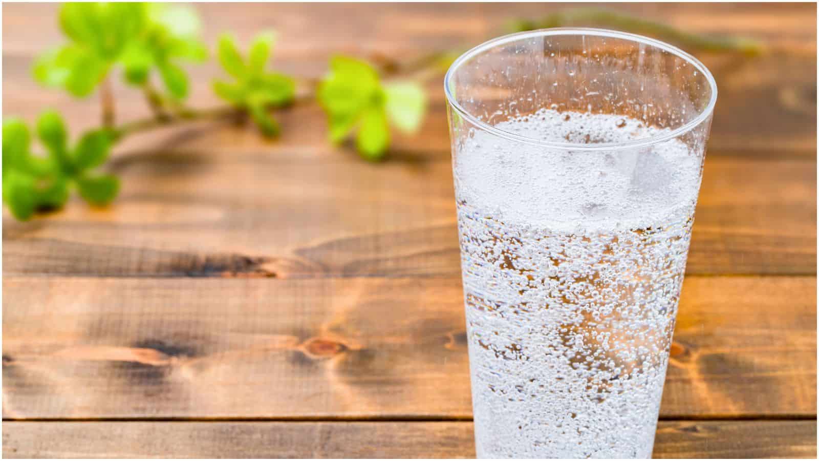 A clear glass filled with sparkling water is placed on a wooden table, standing in contrast to the list of sugary drinks often seen. The background includes green leaves slightly out of focus.