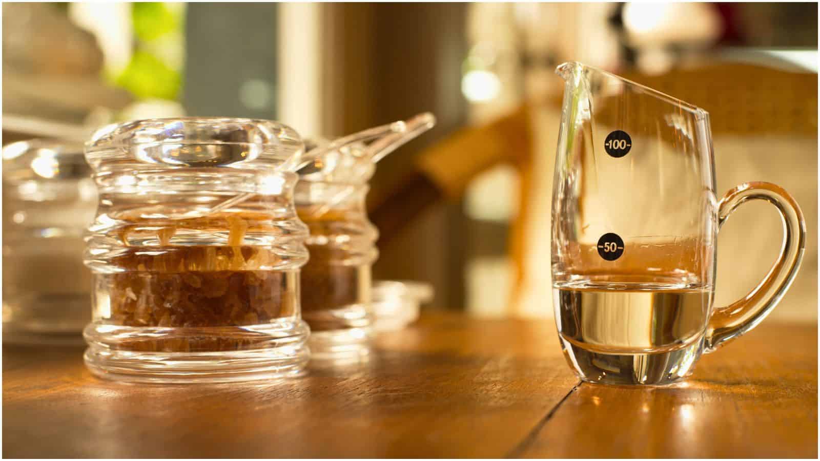 A glass measuring cup filled with liquid and marked with 50 and 100 units is on a wooden surface. Two glass jars containing a brown substance equipped with spoons are in the background, with a blurred indoor setting visible. This setup offers everything you need to know about simple syrup preparation.
