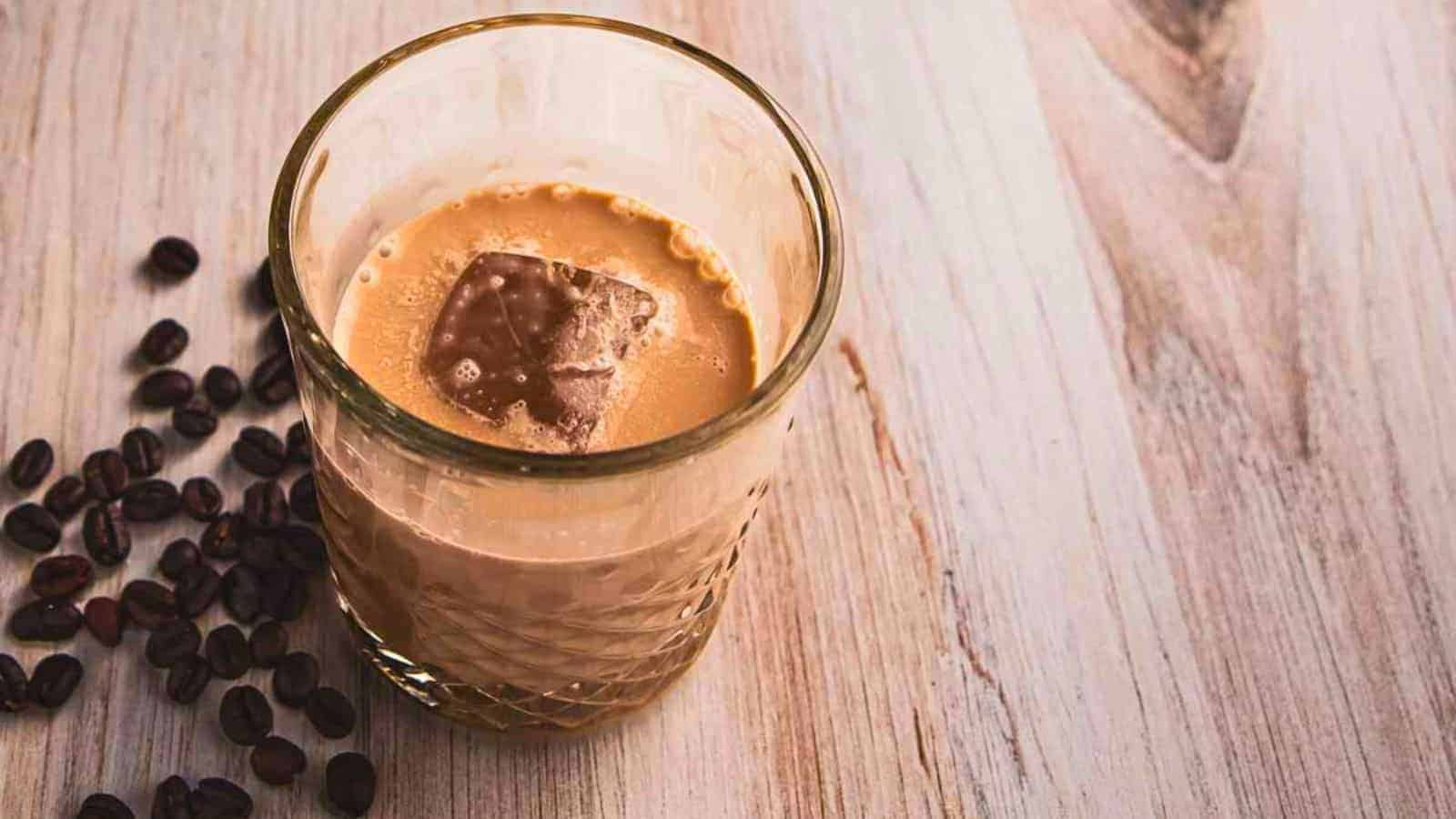 A glass of iced coffee with milk and ice cubes is placed on a wooden surface. Beside the glass, there are scattered coffee beans. The surface has a light, rustic wood texture, offering everything you need to know about simple syrup for your perfect blend.