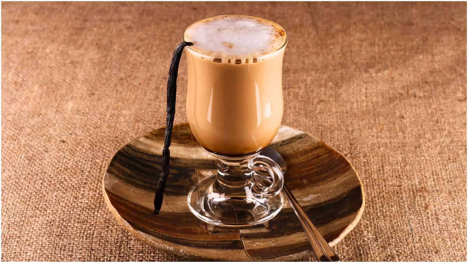 A clear glass mug filled with frothy coffee sits on a round, striped wooden saucer. A stick of vanilla rests against the mug's rim, and a metal spoon lies on the saucer. The background is a textured, brown fabric surface, evoking the warmth of spices for coffee.