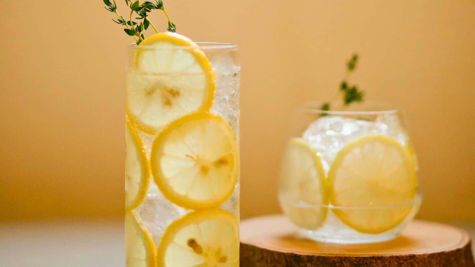 Two drinks with lemon slices and herbs garnish. The drink on the left is in a tall glass, while the one on the right is in a short, round glass, both placed against a light, neutral background. Perfect for enjoying lemon water before bed.