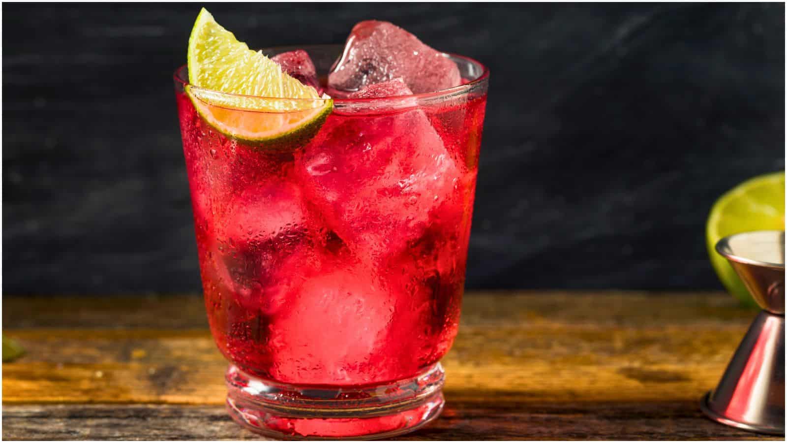 A clear glass filled with a red beverage and large ice cubes, garnished with a wedge of lime, sits on a wooden surface. A metal jigger and a partially visible lime are in the background&mdash;a subtle reminder that some alcoholic drinks that are bad for your teeth can also look enticingly refreshing.