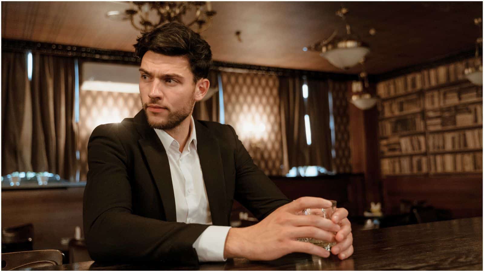 A man with dark hair and a beard is seated indoors at a wooden bar, holding a glass in one hand. Dressed in a white shirt and dark jacket, he looks pensive. The background features dim lighting, chandeliers, dark curtains, and wall-mounted bookshelves, all respecting the establishment's strict alcohol rules.