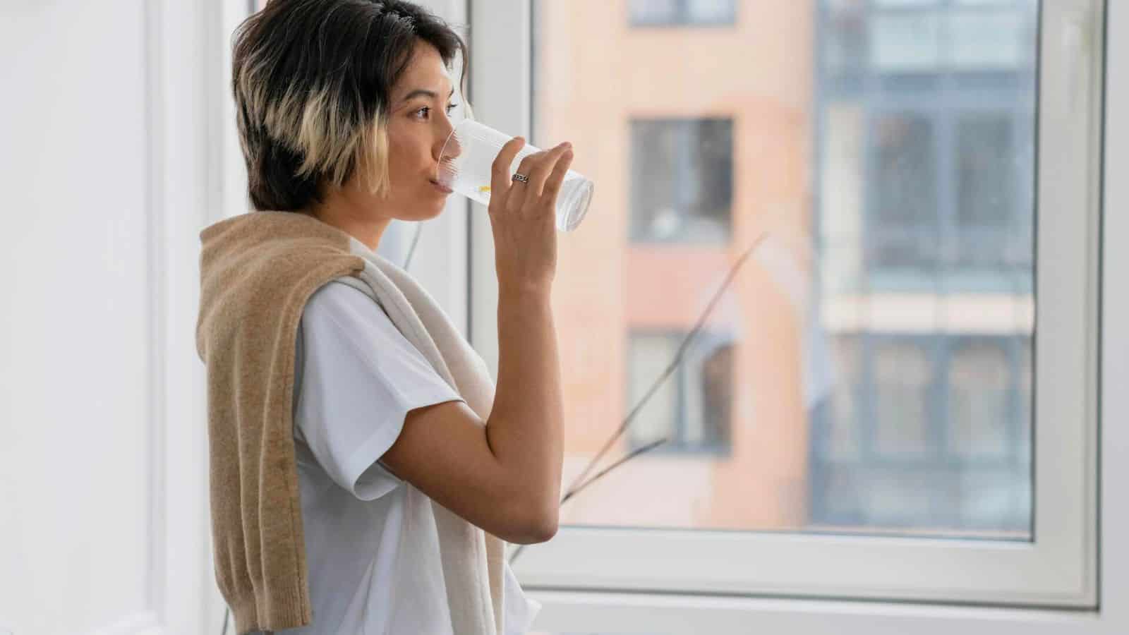 A person with short, dark hair and a light streak on the side is standing by a window, drinking water&mdash;one of the best drinks for gout. They are wearing a white t-shirt with a light brown sweater draped over their shoulders. The background shows a blurred cityscape.