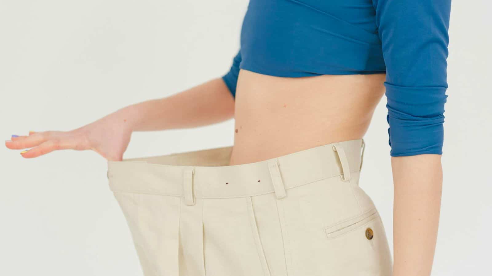 A person wearing a blue crop top shows off a pair of beige pants that appear too large by extending the waistband outward with one hand, showcasing how much they've slimmed down since they decided to stop drinking alcohol.